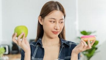 A woman is deep in thought while holding a doughnut in one hand and an apple in the other hand.