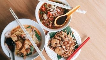 Bowls of curry laksa, assam laksa and a plate of char kuey teow on a wooden table