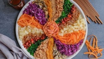 Colourful Chinese New Year yee sang platter on a grey table with a glass of tea, carrot strips, chopsticks, and a kitchen towel.
