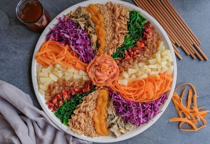 Colourful Chinese New Year yee sang platter on a grey table with a glass of tea, carrot strips, chopsticks, and a kitchen towel.