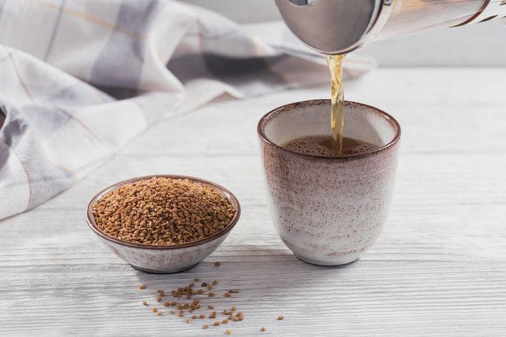 Fenugreek tea is poured into a cup next to a small bowl of fenugreek seeds on a white-painted wooden table.