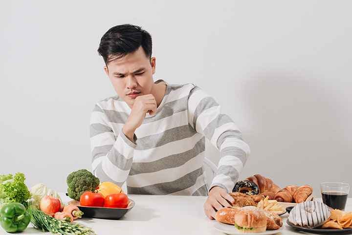 Man pushes away unhealthy high blood pressure foods placed on while table to his left, while fresh vegetables are to his right.