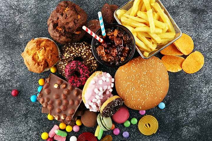 Rich, fried, salty, and sweet foods, including a hamburger, french fries, chocolate, doughnuts, soft drink, and sweets on a grey marbled surface.
