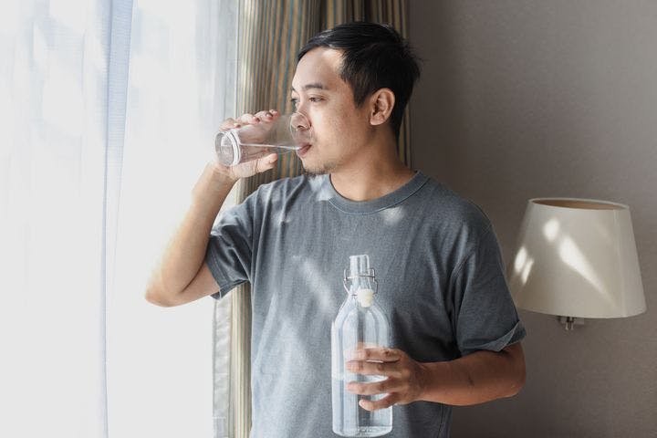 Man drinks a glass of water while looking out the window and holding a bottle of water.