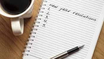 Notebook page reads “new year resolutions” with five blank numbers spaced next to a cup of coffee on a wooden table.