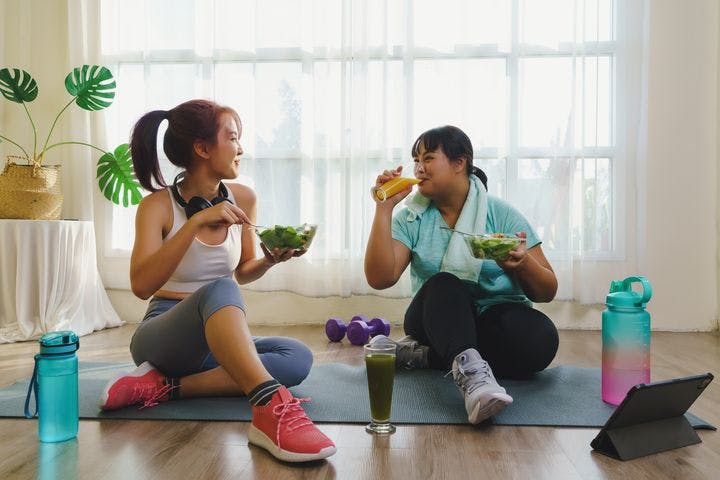Two women smile at each other while in exercise clothes sitting on a yoga mat eating healthy food.