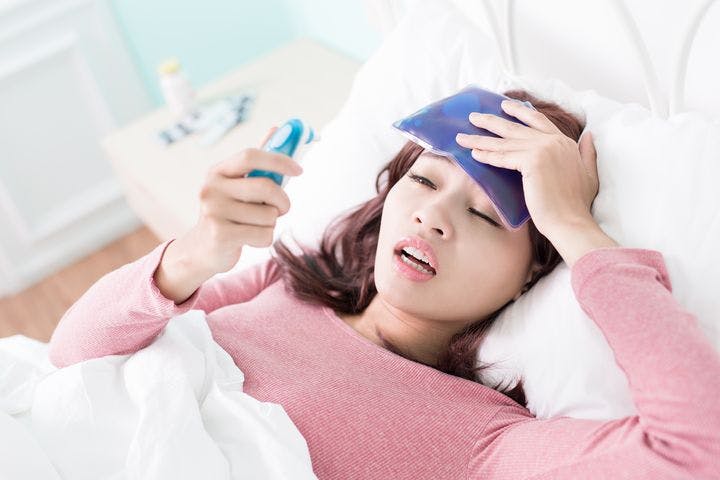 Sick woman in bed holding a cooling patch to her forehead with her left hand while looking at an ear thermometer.