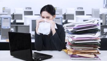 Woman in an office sits next to a tall pile of work folders while holding a tissue over her runny nose.