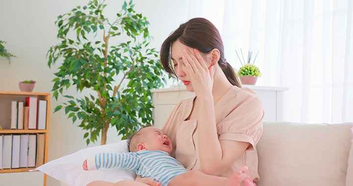 New mother holds a crying baby while holding her head and closing her eyes with a tired expression.