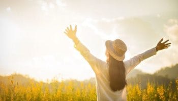 Woman wearing a hat outside in a field of yellow spring flowers raises her arms, welcoming the spring sun.