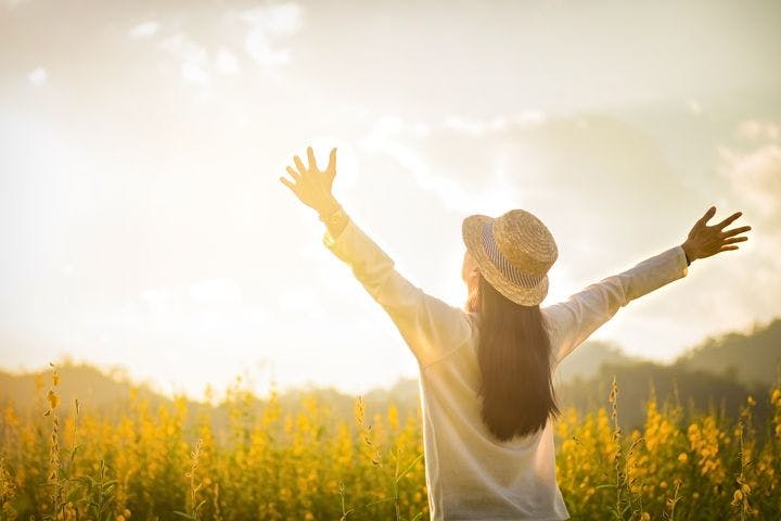 Woman wearing a hat outside in a field of yellow spring flowers raises her arms, welcoming the spring sun.