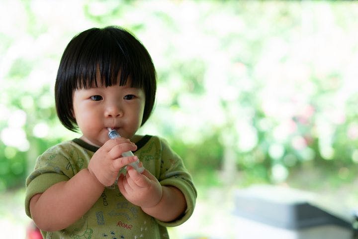 A toddler girl takes oral medicine by herself through a syringe she holds with her hands.