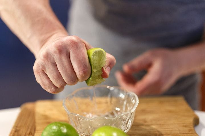 Man squeezing juice from a lime wedge into a glass bowl placed on a wooden cutting board.
