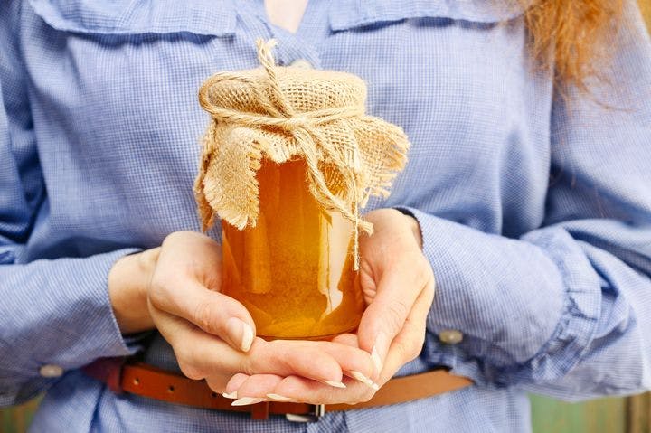 Woman using both hands to hold a glass jar of honey that’s covered with a rattan cloth and string.