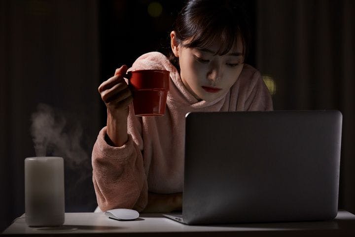 Woman holding a coffee mug in her right hand while looking at a laptop and sitting at a desk with a humidifier.