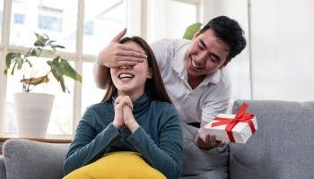Smiling man closing a woman’s eyes while holding a gift box in his left hand as she reacts with surprise while sitting on a sofa.