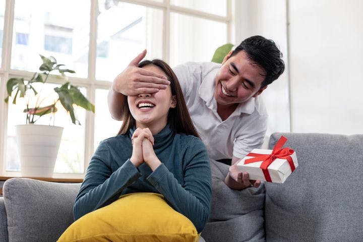 Smiling man closing a woman’s eyes while holding a gift box in his left hand as she reacts with surprise while sitting on a sofa.
