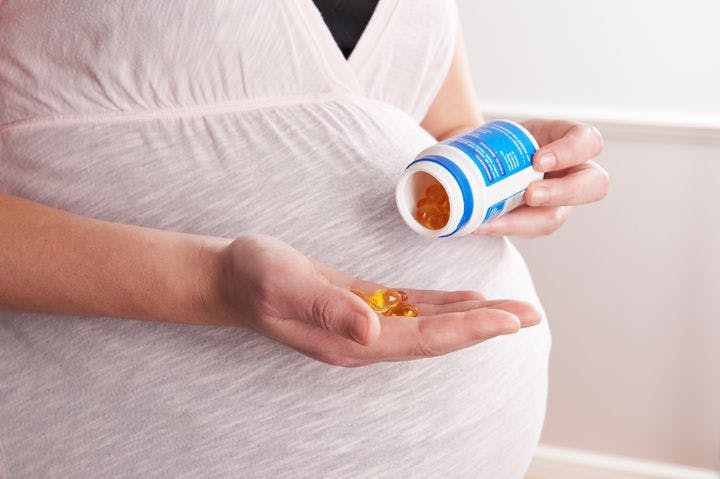 Pregnant woman uses her left hand to drop cod liver oil supplements from a plastic bottle into her right palm.  
