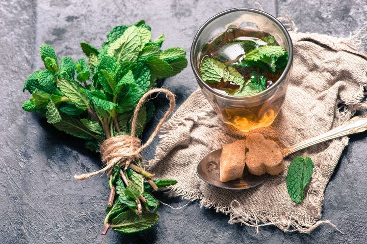 Cup of tea garnished with peppermint leaves, a spoonful of sugar on a jute fabric, and peppermint leaves tied together.