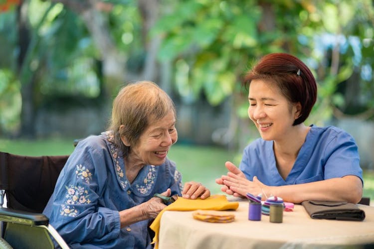 Elderly woman in a wheelchair and her caregiver smiling together as they work on a craft project together while sitting at a table.