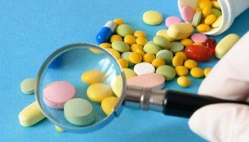 Pills and supplements coming out of a bottle strewn against a blue background with a gloved hand holding a magnifying glass in the foreground.