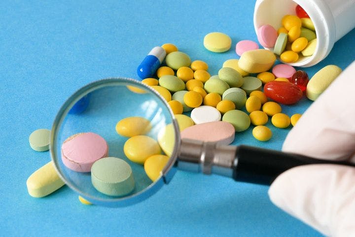 Pills and supplements coming out of a bottle strewn against a blue background with a gloved hand holding a magnifying glass in the foreground.