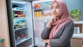 Woman with a thinking expression stands by a fully stocked fridge with the door open.