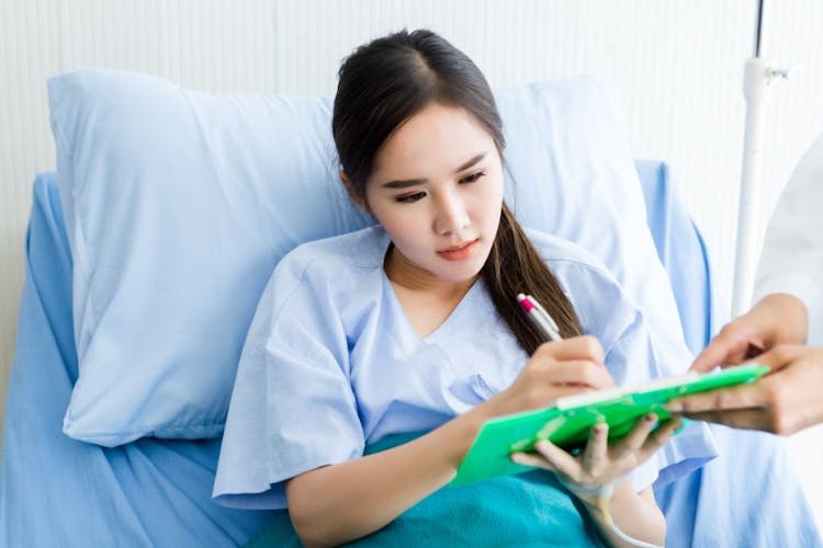 Woman in patient gown signing a consent form while sitting up in bed