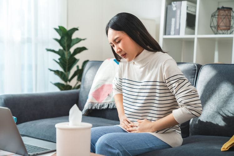 An Asian woman holding her stomach in pain while sitting on the couch. 
