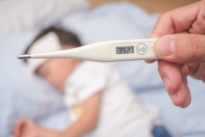Close-up image of a thermometer with a reading of 39.2 degrees Celsius (˚C) as a boy is seen lying down with a fever patch on his forehead.