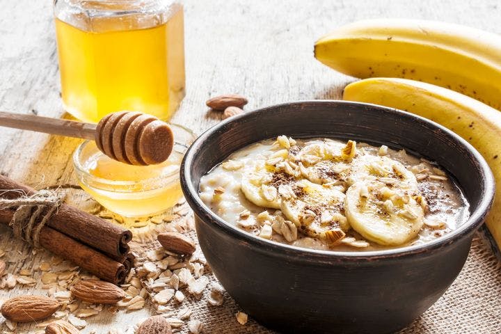 Close-up of oatmeal porridge with chopped nuts and bananas in a black-coloured bowl alongside honey, almonds, bananas and cinnamon sticks on a wooden table. 
