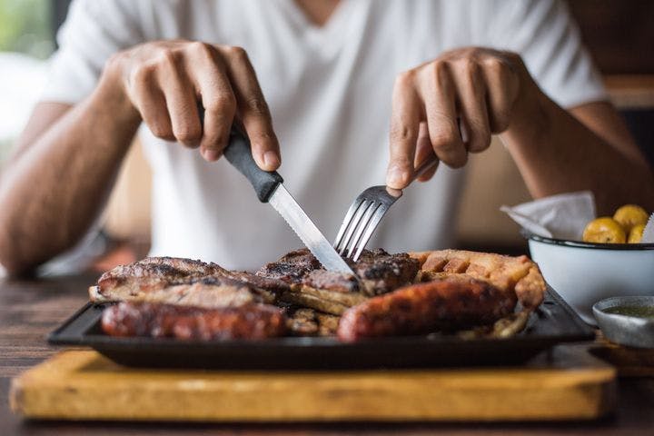Man using fork and knife to cut into a steak on his plate which also has other cooked meats.