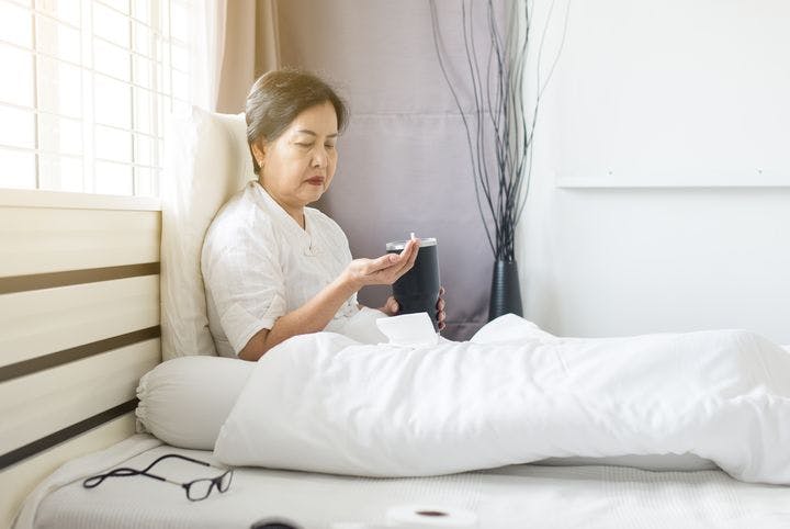 Older woman sits in bed holding a flask in one hand and pills in the other hand.