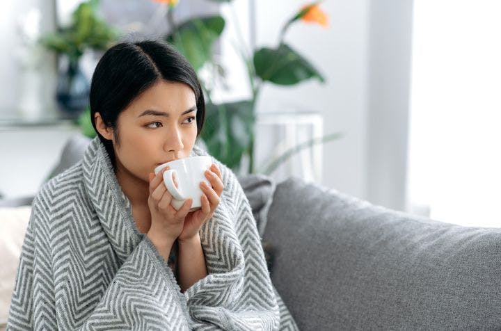 A woman wrapped in a blanket sipping a beverage from a mug she holds with both hands while sitting on a sofa.