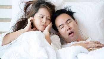 Disgruntled woman closing her ears while looking at a man who’s sleeping with a wide-opened mouth as they both lie in bed.
