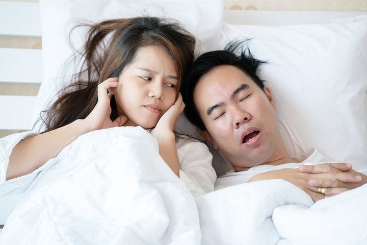 Disgruntled woman closing her ears while looking at a man who’s sleeping with a wide-opened mouth as they both lie in bed.