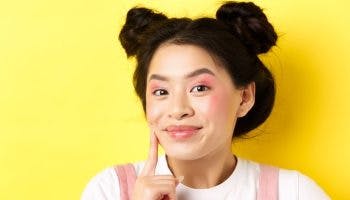 Young hip girl with a rosy complexion and pink makeup places her index finger on her cheek while smiling at the camera.