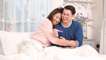 Couple embrace joyfully as they look at a pregnancy test kit in bed.