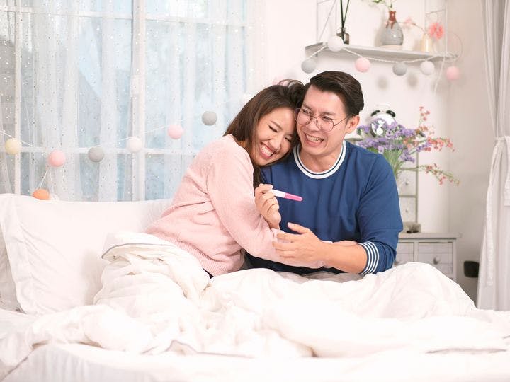 Couple embrace joyfully as they look at a pregnancy test kit in bed.