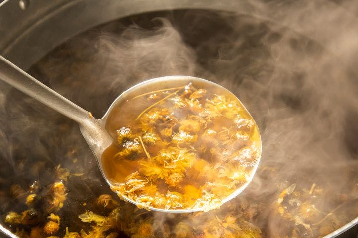 Ladle filled with chrysanthemum flowers boiled in hot water; a large cooking pot of the same tea in the background.