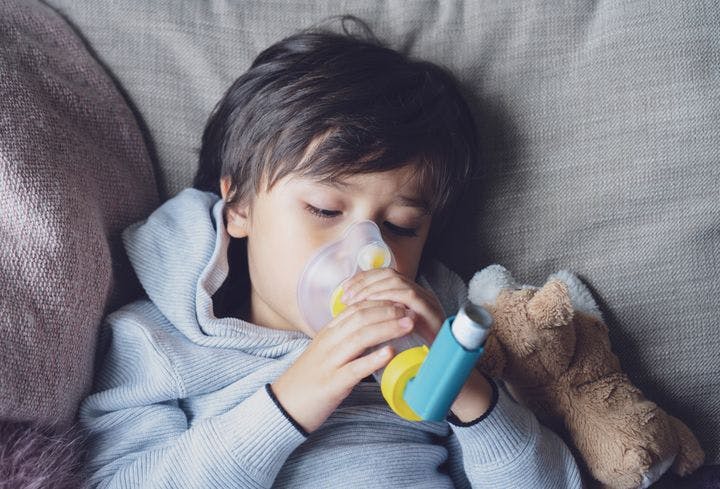 Boy holding an asthma pump with a spacer to his mouth and nose while leaning back into a sofa next to a plush toy. 