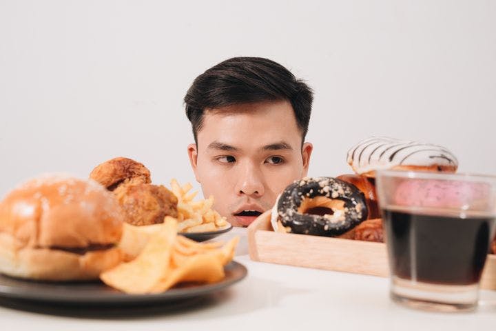 Man stares at a table filled with rich, sweet, salty foods including a soft drink, donuts, hamburger, fried chicken, fries, and chips.