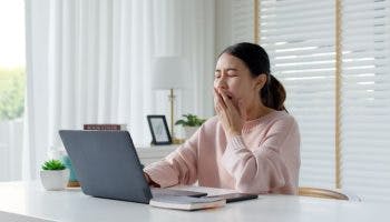 Woman yawning while covering her mouth with her left hand as she sits at a desk in front of a desktop computer.