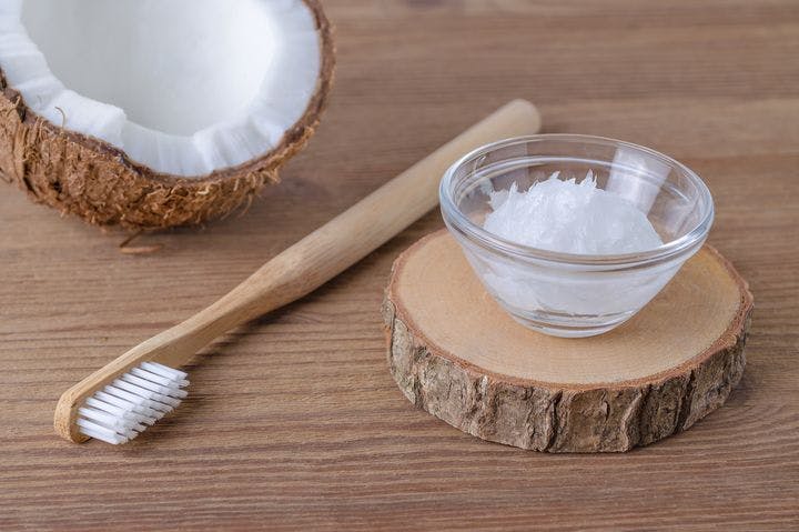 An opened coconut, coconut oil toothpaste, and a wooden toothbrush on a wooden slab.     