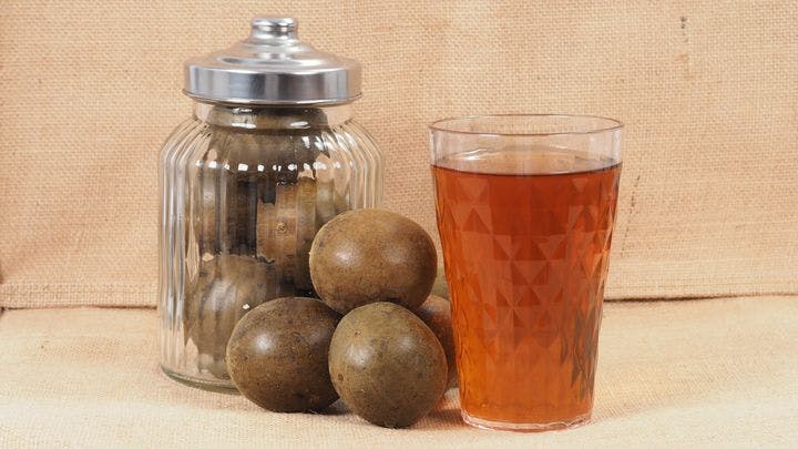 Close-up of dried monk fruits in a closed-lid glass jar and on a table next to a glass of monk fruit tea. 