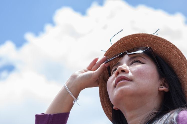A woman lifts her sunglasses with her right hand as she looks up to the sky.