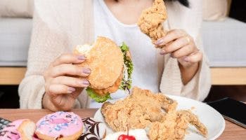 A woman holds a piece of fried chicken and a burger with more food on a table.