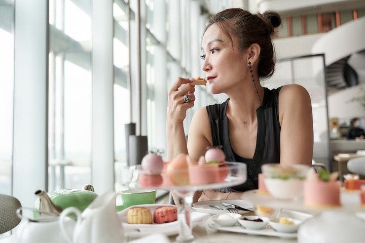 A woman holding a pastry while looking out a window, sitting at a table with pastries and other desserts. 