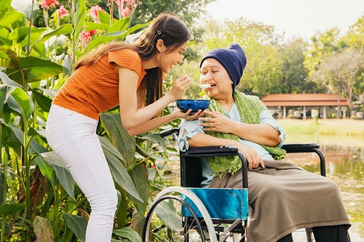 Younger woman spoon-feeds older woman in wheelchair outside.