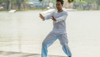 Man wearing white long-sleeved shirt and pants practices Qigong on a yoga mat next to a lake.